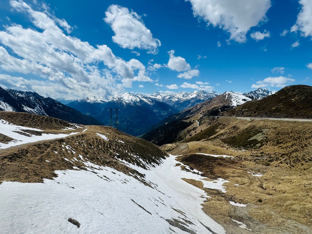 Panoramic view from an Alpine pass summit showing snow patches on mountain slopes and a winding road below with snow-capped peaks in the distance under a vivid blue sky