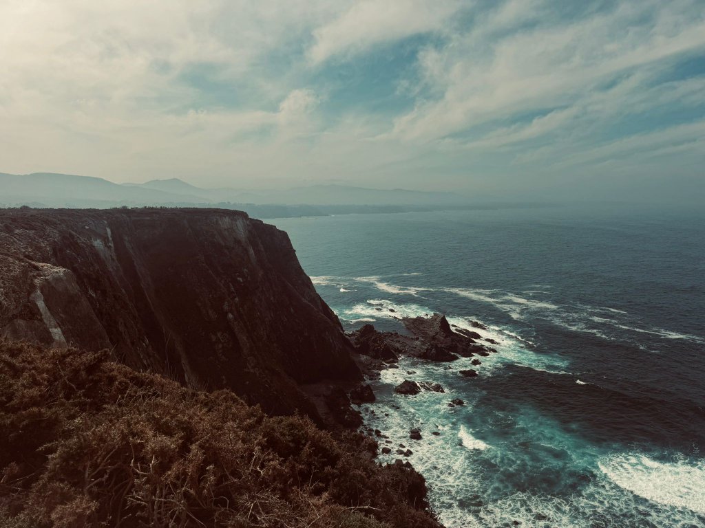 Dramatic Atlantic coastline cliffs dropping vertically into a deep blue sea with waves breaking at the base and mountains visible in the distance under a moody sky