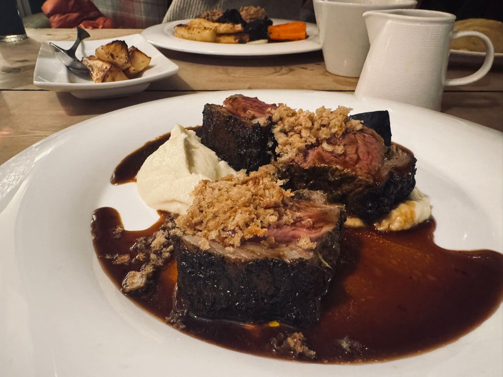 Beautifully presented slow-braised beef with rich dark sauce, whipped potato and breadcrumb crust served at a rustic wooden table, with other plates of roasted vegetables visible in the background