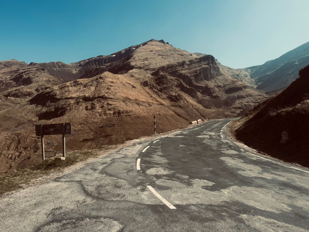 Empty high mountain pass road winding into the distance with dramatic bare rocky peaks under a clear blue sky and a route marker sign visible on the left