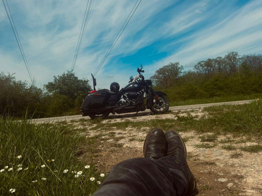 Motorcycle rider lying back in the grass beside a country road with a black Harley Davidson Road King parked nearby, boots visible in the foreground, daisies and blue sky above