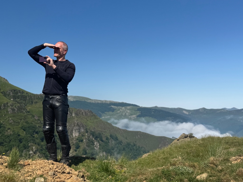 Wild Roads rider shielding eyes photographing cloud inversion — Pyrenean summit — May 2026