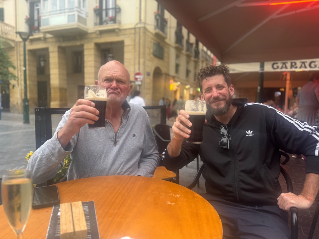 Two Wild Roads riders raising pints of Guinness at pavement cafe — San Sebastian old town — May 2026