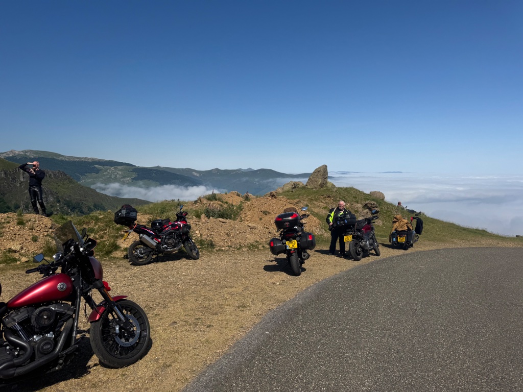 Wild Roads bike fleet parked above cloud inversion on rocky Pyrenean pass — red Harley Sportster foreground — May 2026