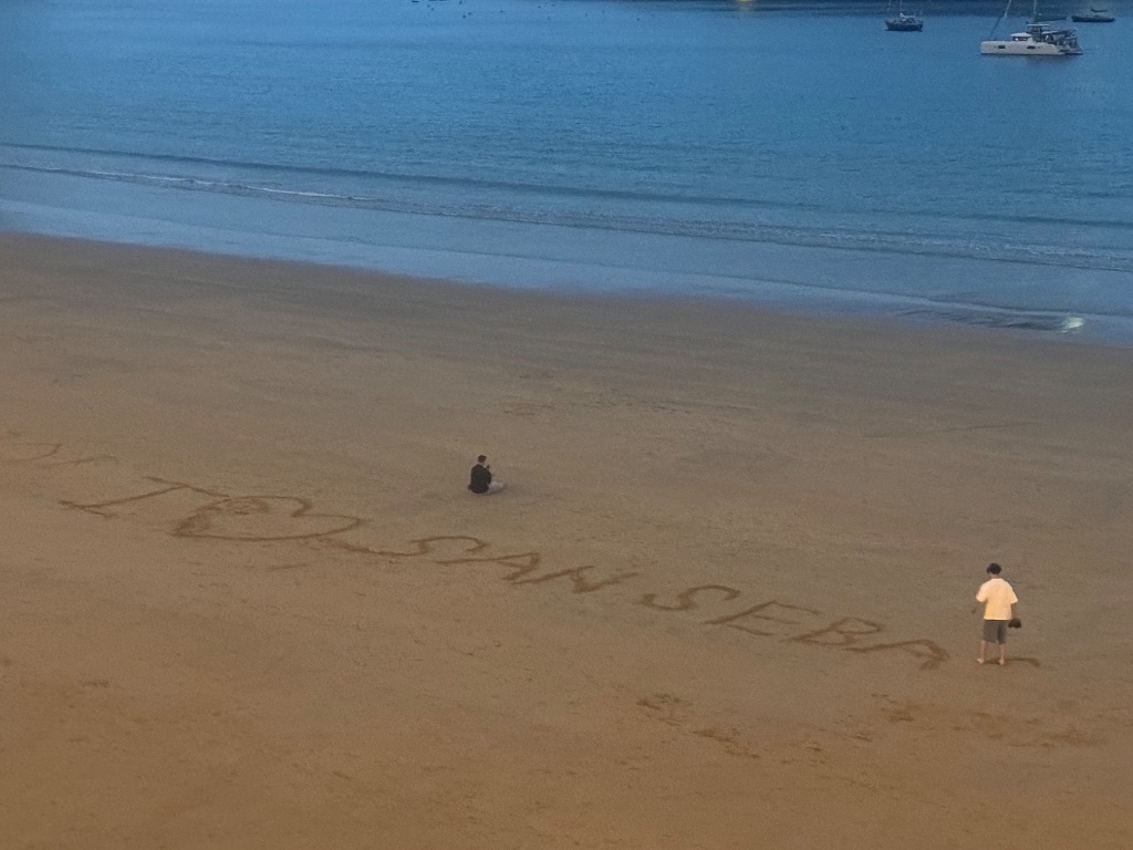 I love San Seba written in sand on La Concha beach at dusk — sailboat in bay — Wild Roads May 2026
