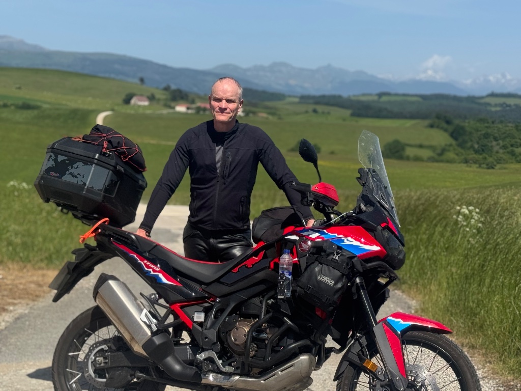 Rider with red Honda Africa Twin on Pyrenean mountain track — snow-capped peaks behind — Wild Roads May 2026