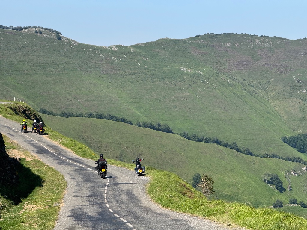 Four Wild Roads riders on narrow twisting road through Pyrenean hillside — green mountains — May 2026