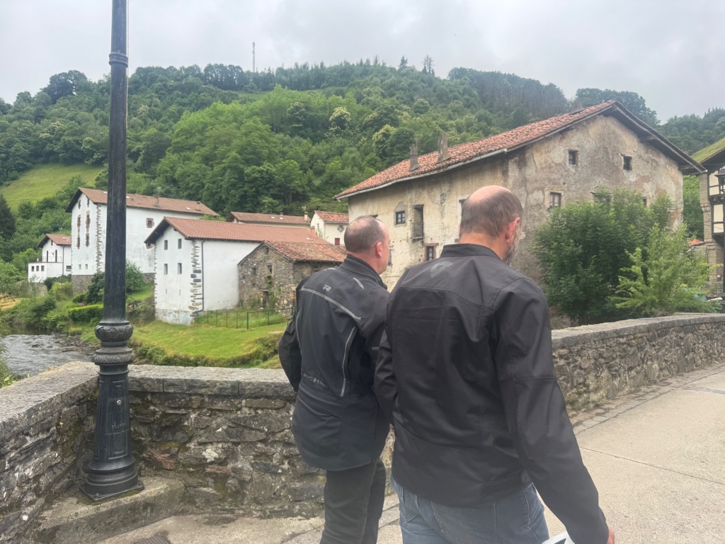 Two riders on stone bridge overlooking Basque village river — Picos & Pyrenees May 2026