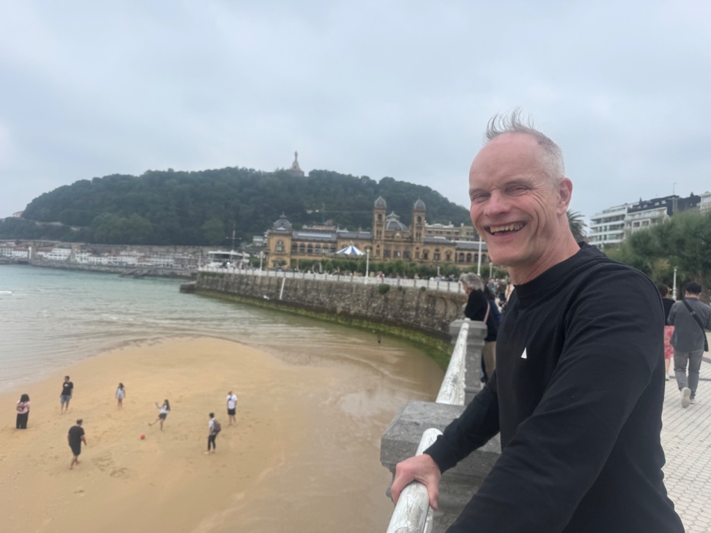 Rider laughing on La Concha beach promenade — San Sebastian town hall and Monte Urgull behind — Wild Roads May 2026