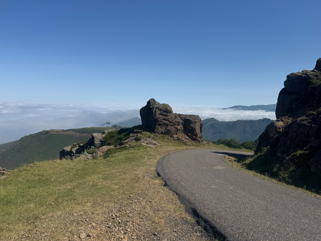 Narrow mountain road curving around rock formation above sea of cloud — Pyrenees — Wild Roads May 2026