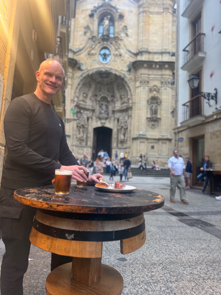 Wild Roads rider with pintxos and beer outside bar in San Sebastian Parte Vieja — Basilica behind — May 2026