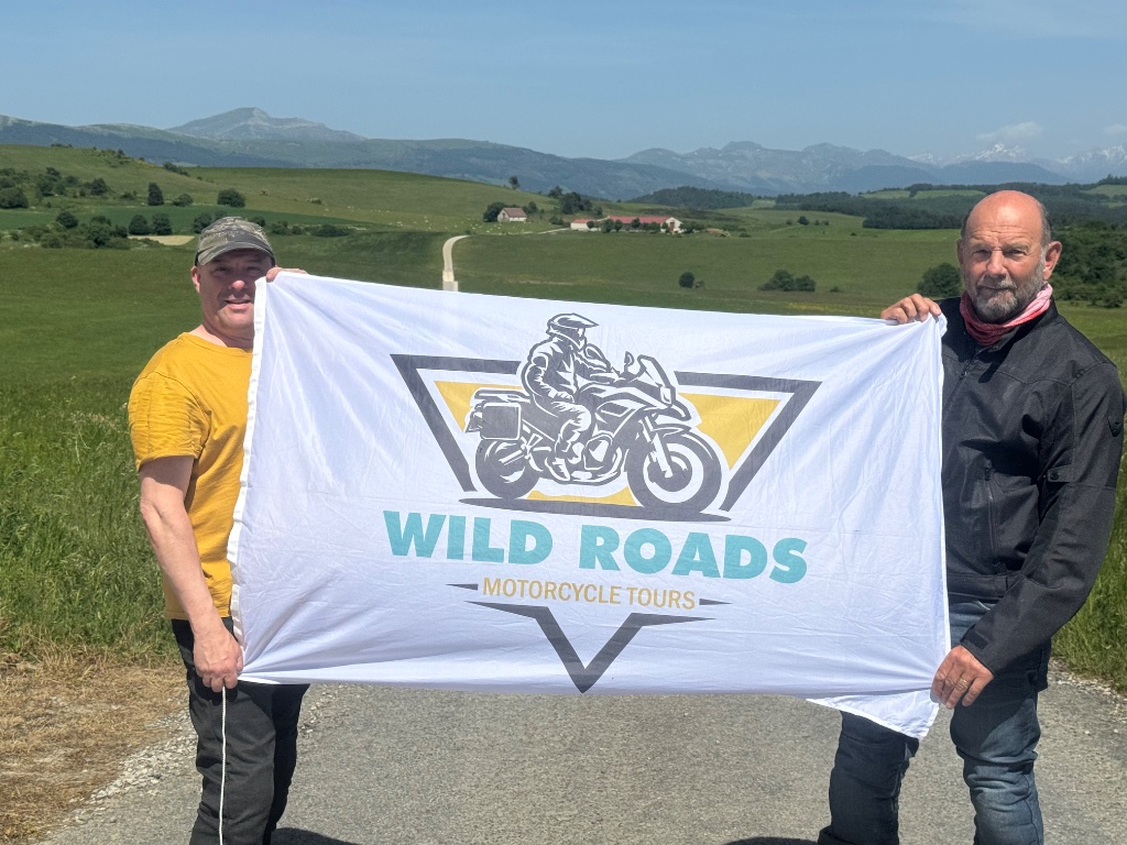 Two riders holding Wild Roads Motorcycle Tours banner on mountain road — Pyrenees foothills — May 2026